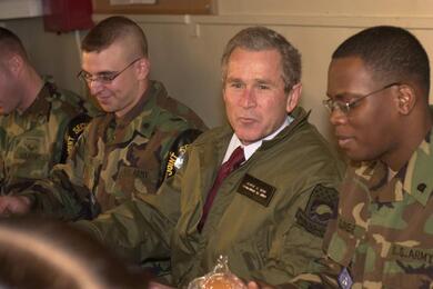 Then-President George Bush in the center-right of the photo while troops sit on either side of him.