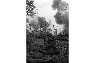 A cavalryman crouches down as smoke billows from North Vietnamese positions hit by artillery in the background.