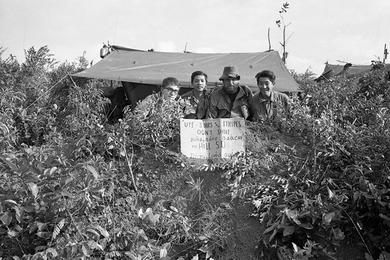 Stars and Stripes photographer John Olson, an unidentified man, UPI photographer William 'Bill' Hall, and UPI photographer Rikio Imajo (left to right) pose behind their tent on which they hung a sign reading "UPI, Stars and Stripes, Don't Shoot" in both English and Vietnamese.