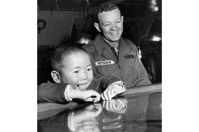 Toddler Hang Nam Chong' chins up to the bar at the Top Three Club at Ascom City as the U.S. Army Signal Depot sponsored a year-end party for orphans of the Dong Chin home which the unit supports. Delighted bystander is PFC Jimmy A. Pettigrew of the 55th Signal Co.