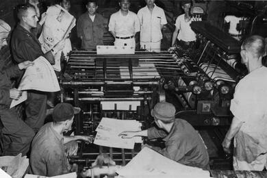 A black and white photo of men in a printing room, some of them examining newspapers.