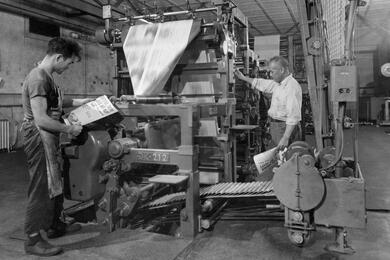 A black and white photo of two men by a large machine in a shop floor, with one man examining a newspaper.