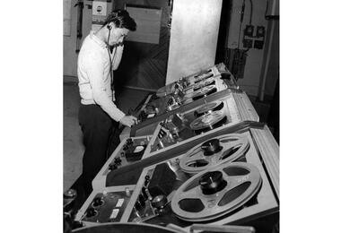 A black-and-white, 1960s-era image of a Japanese man wearing headphones and standing over a large radio control panel.