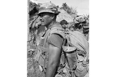 A black and white, vertical photo of a service member in a shirt, with a hut behind him.