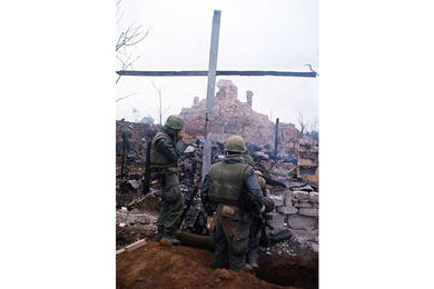 A U.S. Marine radios from under a cross formed by support beams of a structure long reduced to rubble during the Tet Offensive. Dong Ba gate and its tower lie amid smoke and ruins in the background.