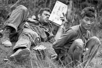 A 1st Cavalry soldier catches up on his reading while a suspected Viet Cong awaits evacuation for interrogation during an operation in the Que Son Valle