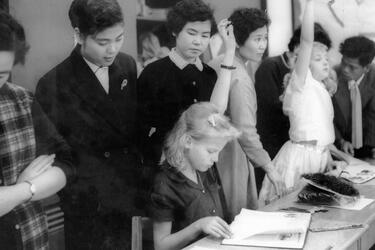 Miss Haruko Ota, a third grade teacher, peeks over the shoulder of Brenda Sheets, 10, daughter of Master Sgt. and Mrs. W. G. Sheets,