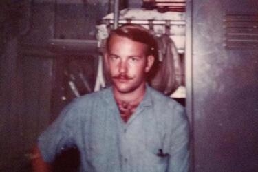A veteran with a mustache and blue shirt stands in front of a brown background.