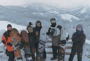 Five men holding snowboards pose for a picture in front of some mountains.